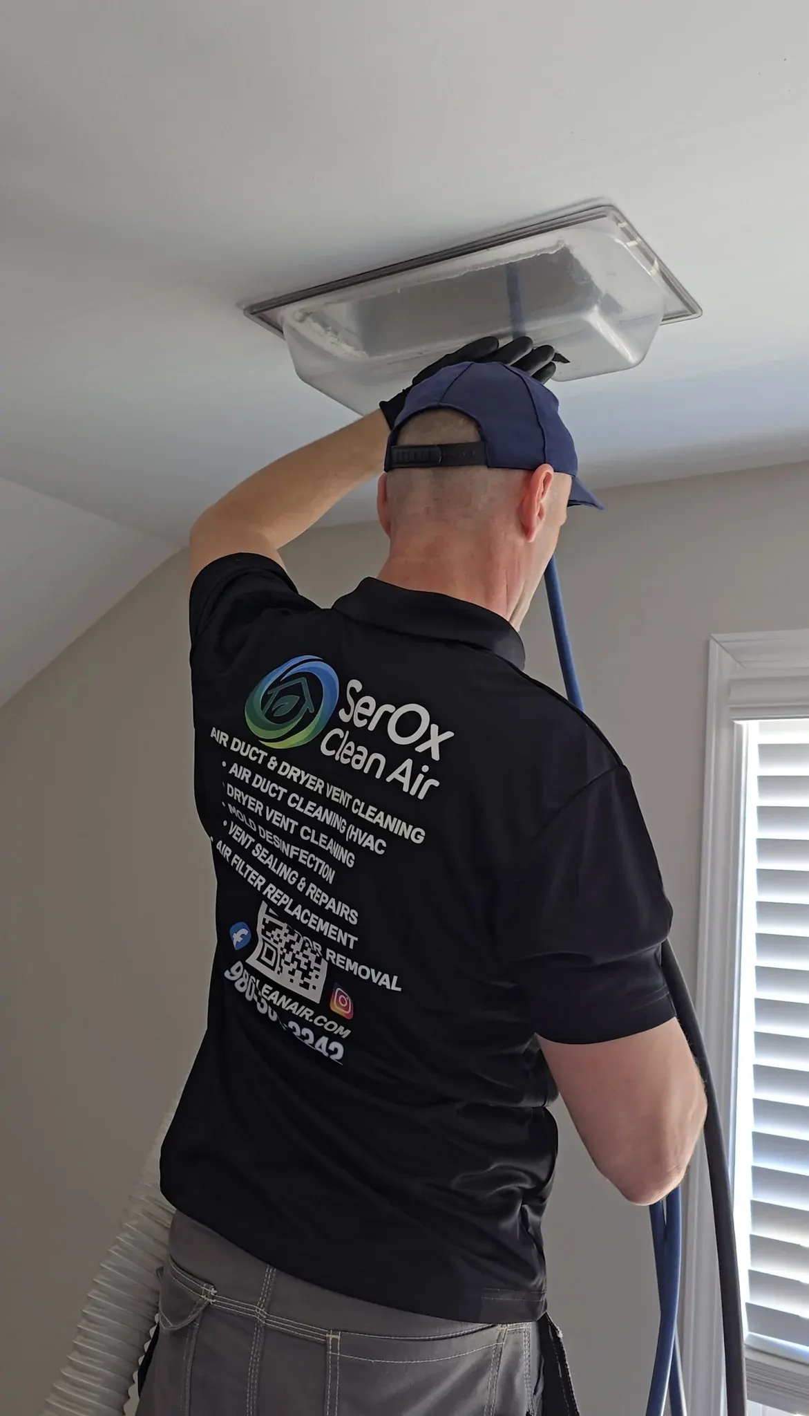 Serhii from behind in a black SerOx polo, cleaning a ceiling vent under a clear dust shield