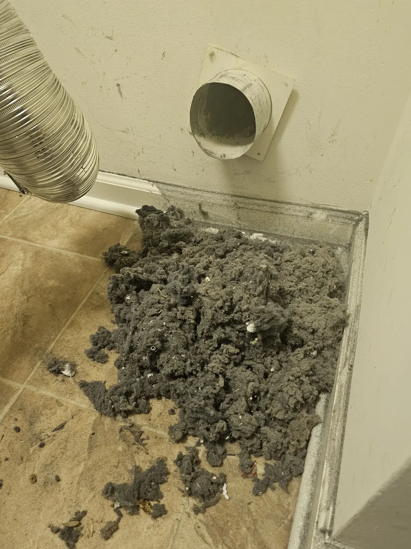 Large pile of lint on the floor next to a dryer vent outlet and hose