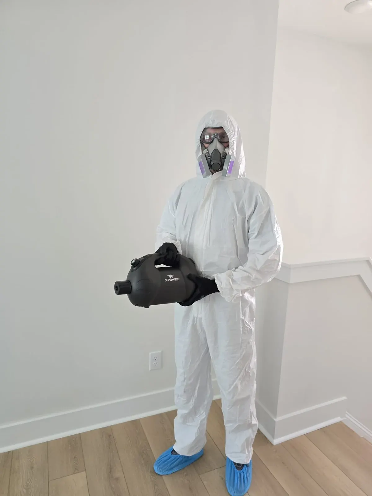 Technician in a protective suit and shoe covers holding a fogger in an empty room