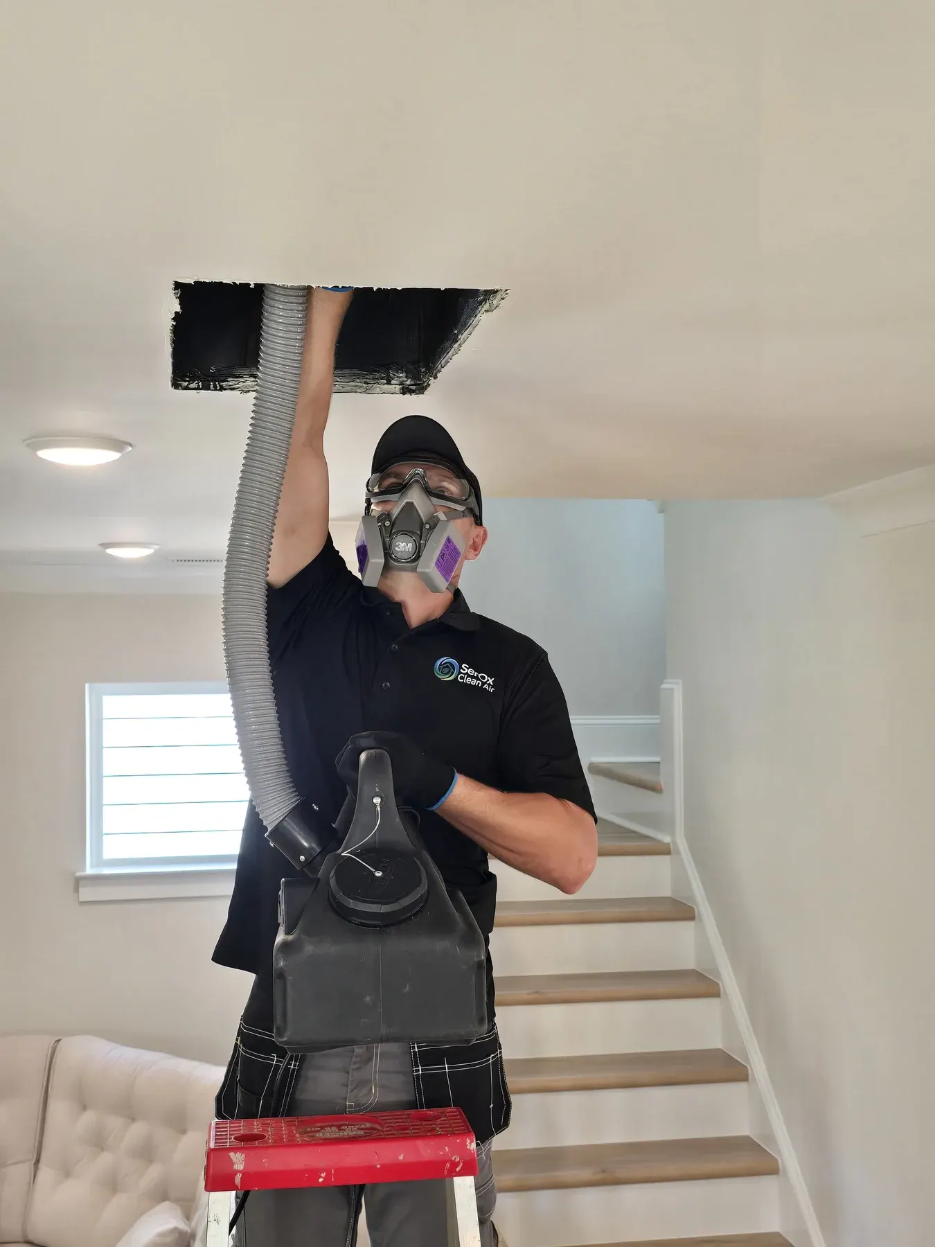 Technician in a black SerOx polo and respirator feeding a vacuum hose into an open ceiling duct