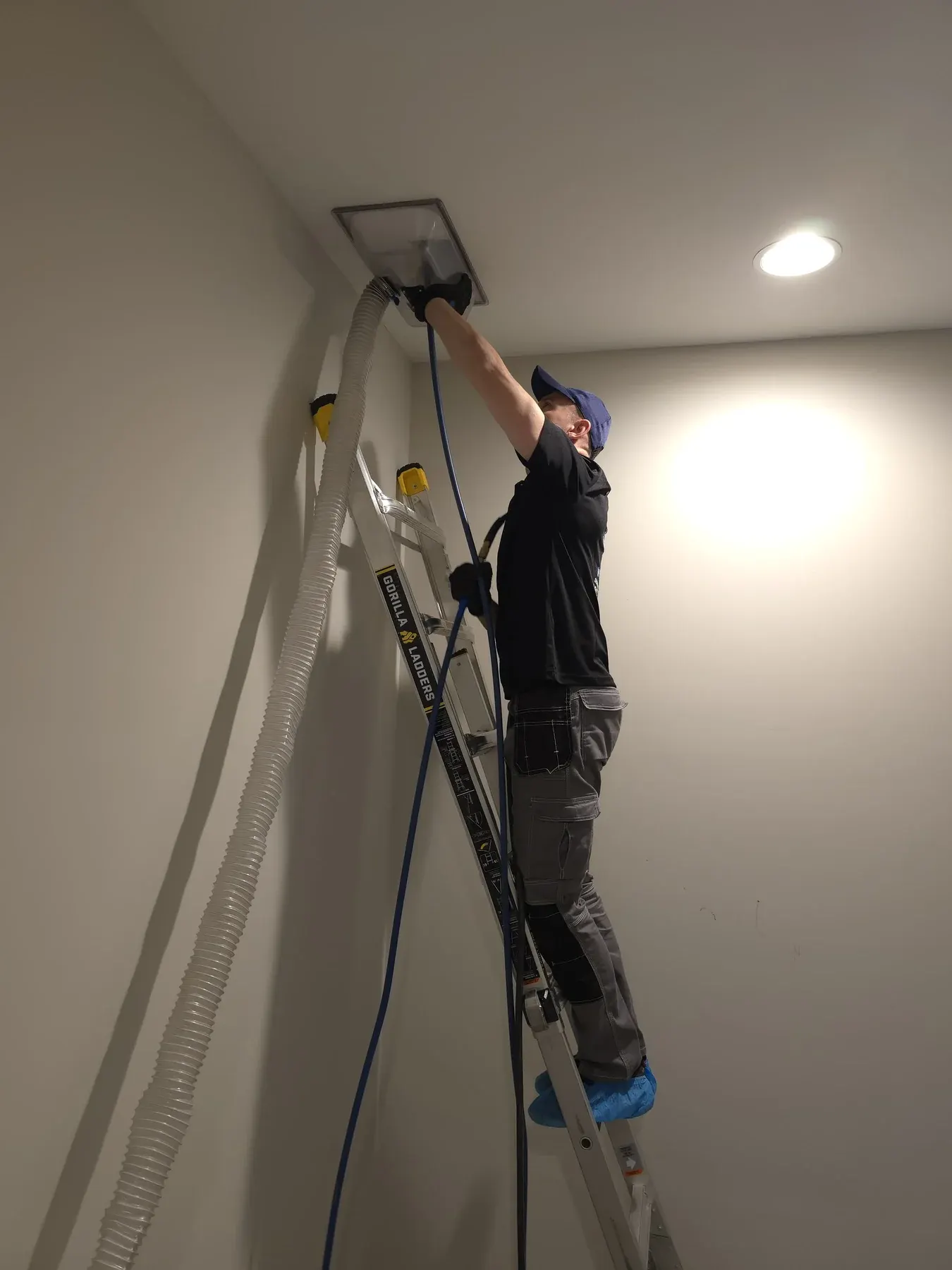 Serhii on a ladder vacuuming a ceiling vent in a hallway